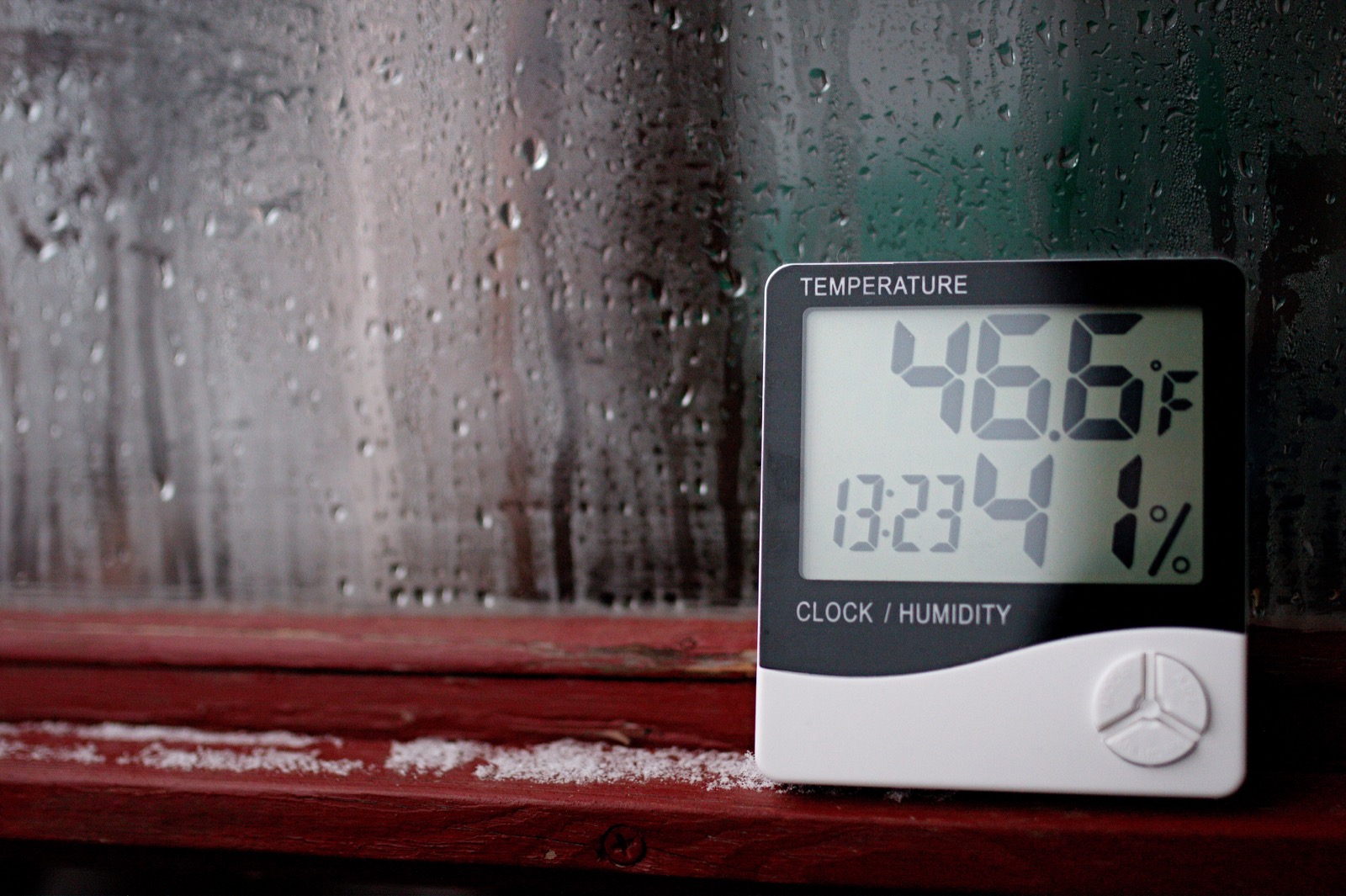 A digital hygrometer displaying indoor temperature and relative humidity beside a condensation-covered window