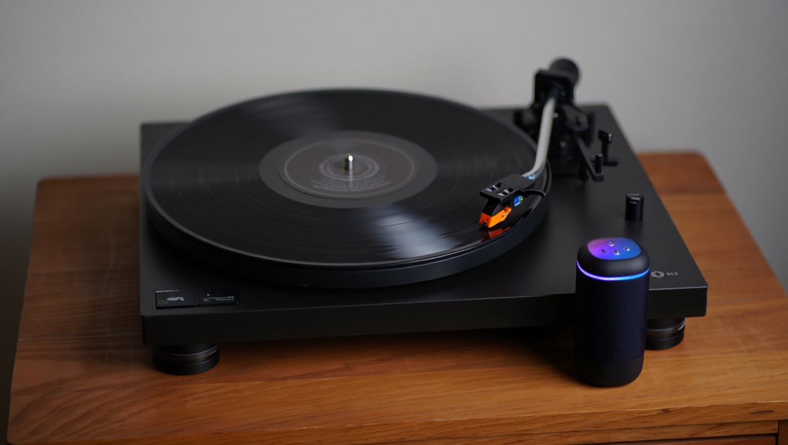 A turntable playing a vinyl record beside a wireless speaker on a wooden sideboard